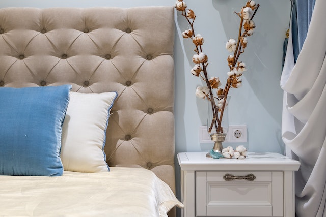 Blue and beige pillows on a bad with flowers in a vase on a nightstand