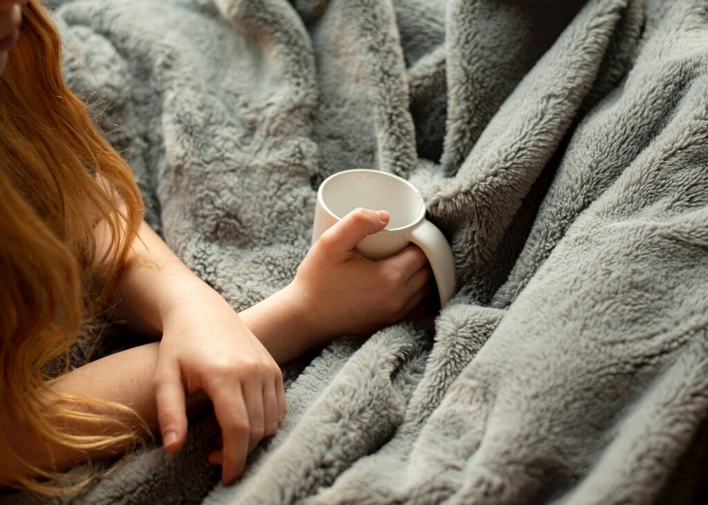 A woman covered with a grey blanket holding a white cup in her hands