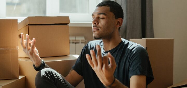 Man meditating between boxes and thinking about creating tranquil spaces with mindful decor after moving.